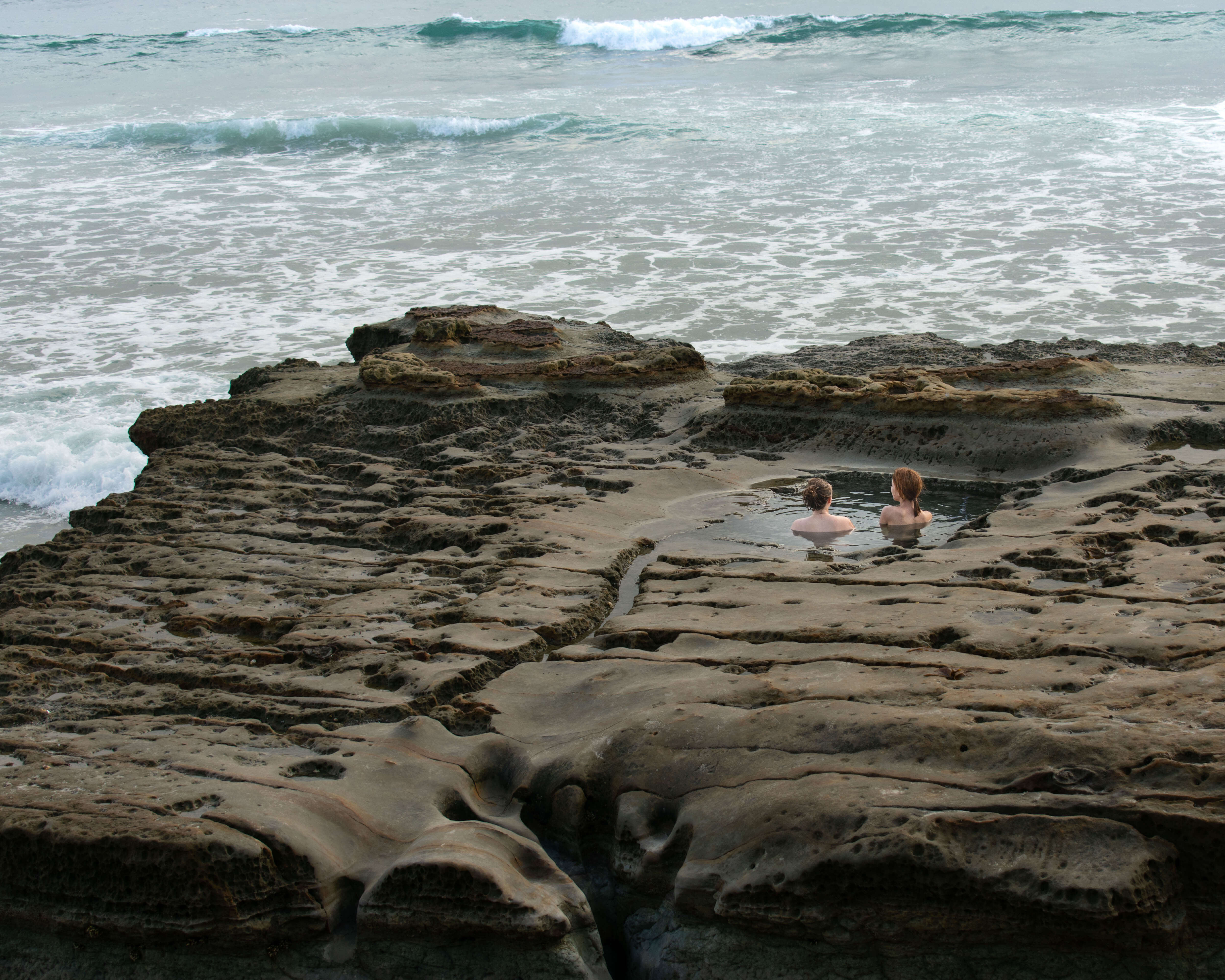 two ladies sit in a natural pool inside a rock overlooking the ocean